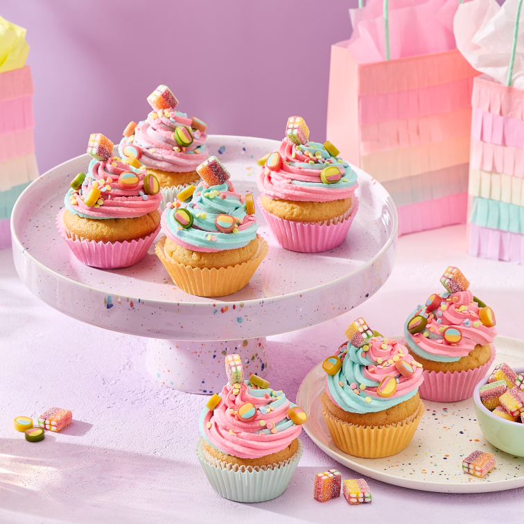 Cupcakes mit rosa und türkisem Frosting in pastellfarbenen Förmchen, dekoriert mit HARIBO Fruchtgummis, angerichtet auf einer gesprenkelten Etagere und einem passenden Teller. Daneben kleine Schälchen mit bunten Süßigkeiten, Gläser mit Getränken und regenbogenfarbene Geschenktüten im Fransen-Look vor einem lila Hintergrund.