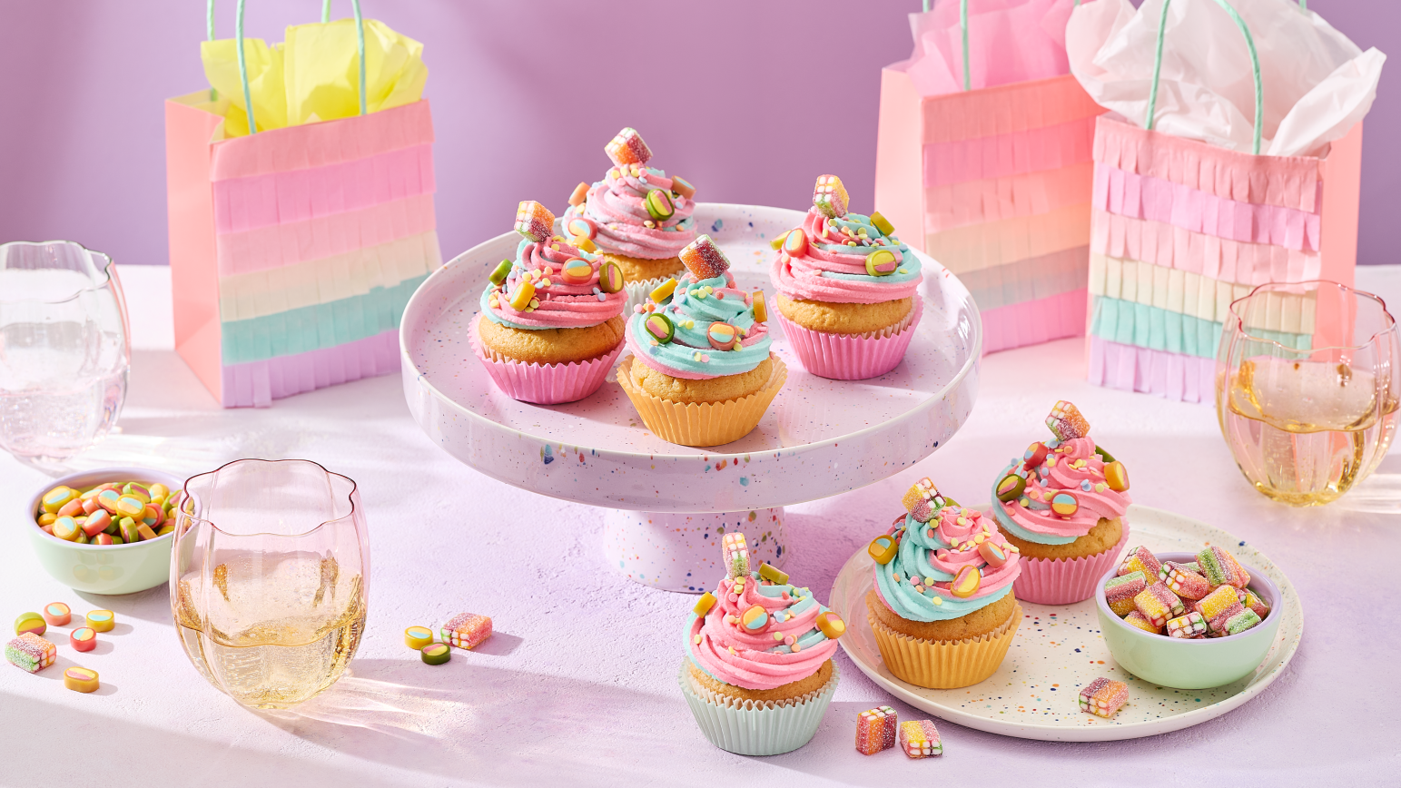 Cupcakes mit rosa und türkisem Frosting in pastellfarbenen Förmchen, dekoriert mit HARIBO Fruchtgummis, angerichtet auf einer gesprenkelten Etagere und einem passenden Teller. Daneben kleine Schälchen mit bunten Süßigkeiten, Gläser mit Getränken und regenbogenfarbene Geschenktüten im Fransen-Look vor einem lila Hintergrund.