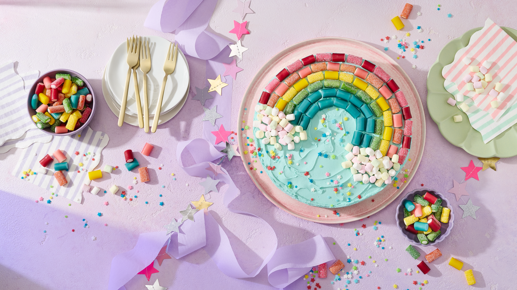 Ein runder Kuchen mit türkisem Frosting, dekoriert mit einem Regenbogen aus HARIBO Pico-Balla, Pico-Balla Sauer, Rainbow und Balla-Balla sowie bunten Pixel-Streuseln auf einem rosa Teller. Daneben Schälchen mit weiteren Fruchtgummis, goldene Gabeln auf weißen Tellern, gestreifte Servietten, eine Sternen-Girlande und lila Satinband auf einem hellflieder Hintergrund – ein farbenfroher Regenbogenkuchen für die Einhorn Party.