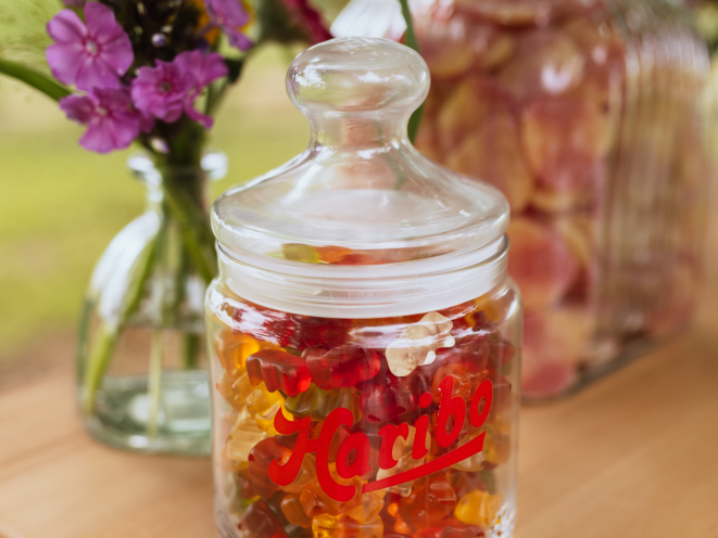An elegant HARIBO-branded glass jar filled with colorful HARIBO Goldbears displayed on a wooden table, with purple flowers in a vase and additional glass jars in the background — a charming wedding candy table setup. The text 'Candybar for the wedding' is displayed in large yellow letters on a blue background, with the subtitle 'The eye-catcher for the big day.' This beautiful and simple arrangement showcases a rustic candy bar wedding look and serves as inspiration for candy for wedding candy bar displays, proving that great candy bar ideas don't have to be complicated.