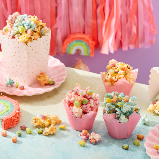 Einhorn Party Essen: Buntes Einhorn-Popcorn in Rosa, Blau und Gelb, serviert in rosa Tulpen-Förmchen und einer weiß-goldenen Popcorn-Box auf muschelförmigen Tellern. Das Popcorn ist getoppt mit HARIBO Pico-Balla, Pico-Balla Sauer, Rainbow, Pixel, Balla-Balla und Chamallows Minis, die auch verstreut auf dem Tisch liegen. Im Hintergrund eine Regenbogen-Piñata, rosa Fransen-Girlande und eine lila Wand – ein kreativer Snack für den Kindergeburtstag.