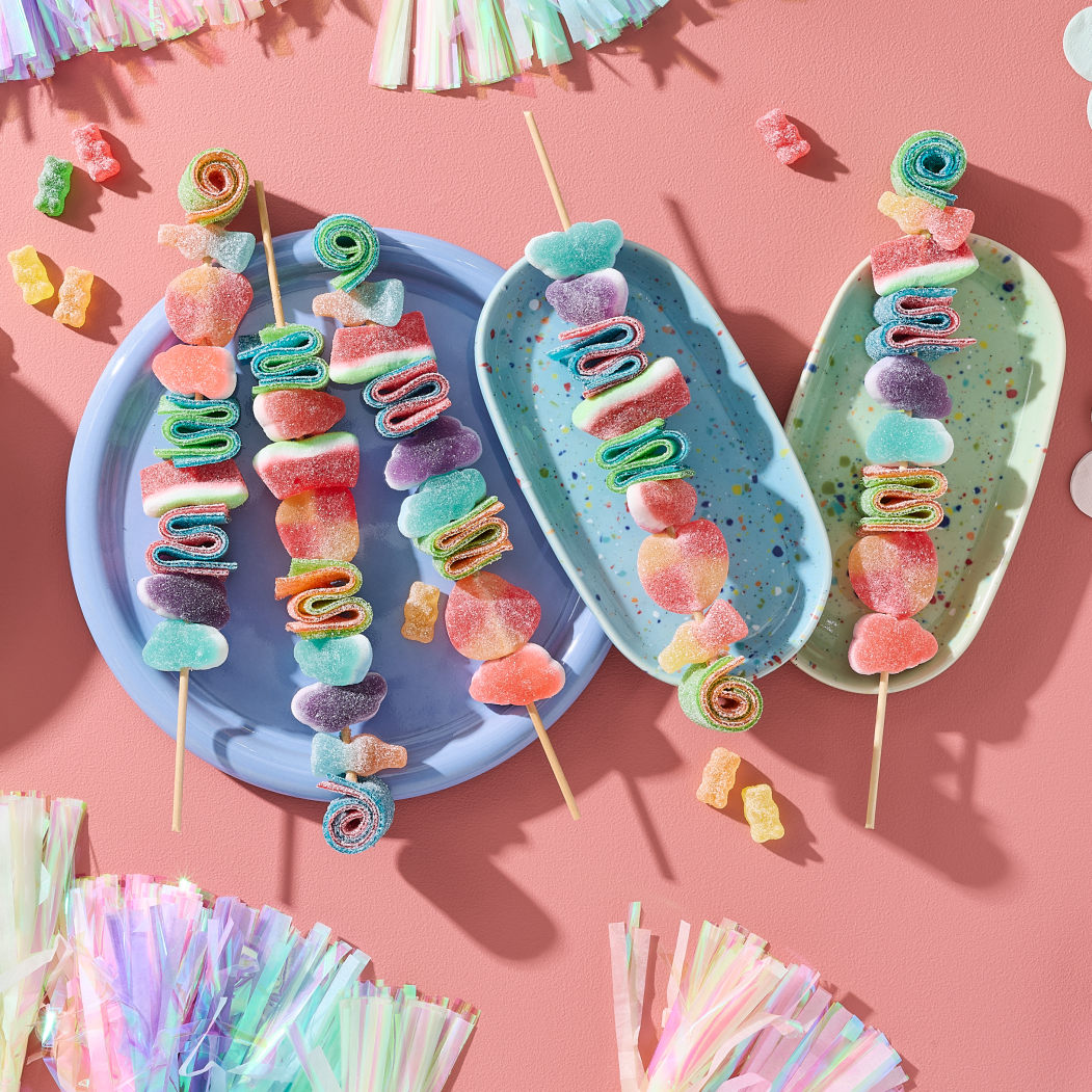 Five colourful candy rainbow skewers loaded with HARIBO gummy sweets including sour sugar-coated hearts, rolled candy spirals and fruity gummy pieces, served on pastel blue and green speckled plates. Next to them is a green bowl filled with more assorted sweet treats. The table is decorated with pastel tassel garlands and cloud-shaped napkins on a coral pink background.