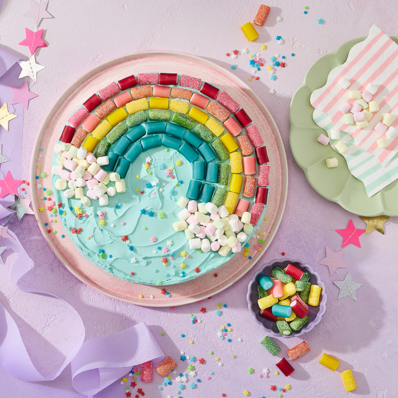Ein runder Kuchen mit türkisem Frosting, dekoriert mit einem Regenbogen aus HARIBO Pico-Balla, Pico-Balla Sauer, Rainbow und Balla-Balla sowie bunten Pixel-Streuseln auf einem rosa Teller. Daneben Schälchen mit weiteren Fruchtgummis, goldene Gabeln auf weißen Tellern, gestreifte Servietten, eine Sternen-Girlande und lila Satinband auf einem hellflieder Hintergrund – ein farbenfroher Regenbogenkuchen für die Einhorn Party.