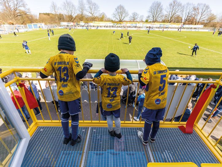 Drei Kinder stehen auf dem Balkon der Pico-Balla-Familienloge und sehen einem Fußballspiel zu.