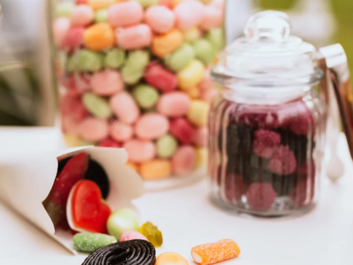 Eine Vielzahl bunter Süßigkeiten in einem Glasbehälter und auf einem Tisch präsentiert. In großen, gelben Buchstaben steht "Snacks", was auf die Auswahl an Snacks für die Geburtstagsparty hinweist. Dieses Bild vermittelt eine süße und festliche Atmosphäre, ideal für Geburtstagsfeier Ideen und die Auswahl von Snacks, um den Geburtstag zu feiern.