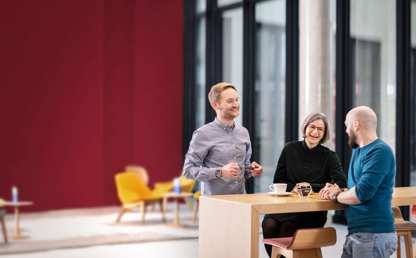 Personen unterhalten sich an einem Tisch mit roter Wand und Fenstern im Hintergrund
