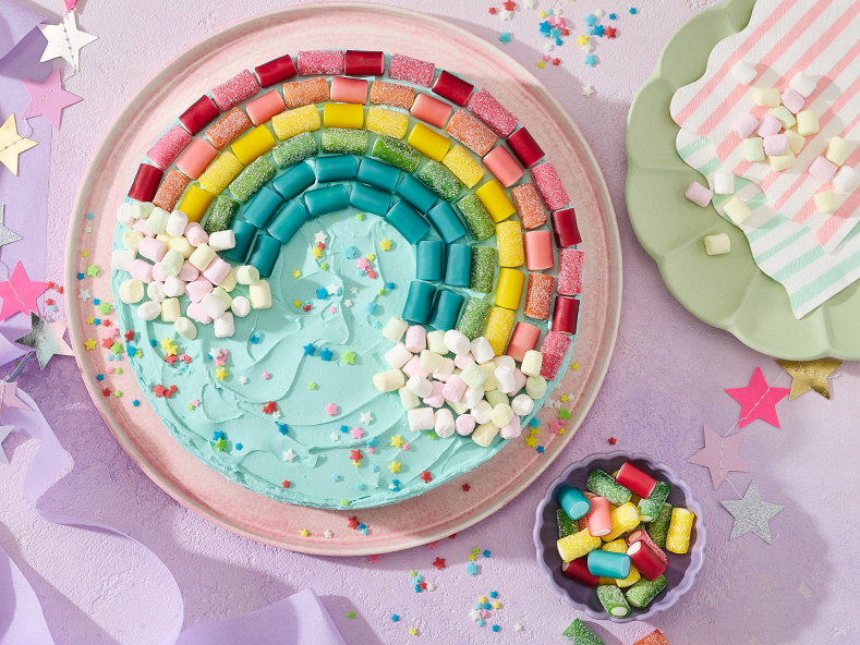 Ein runder Kuchen mit türkisem Frosting, dekoriert mit einem Regenbogen aus HARIBO Pico-Balla, Pico-Balla Sauer, Rainbow und Balla-Balla sowie bunten Pixel-Streuseln auf einem rosa Teller. Daneben Schälchen mit weiteren Fruchtgummis, goldene Gabeln auf weißen Tellern, gestreifte Servietten, eine Sternen-Girlande und lila Satinband auf einem hellflieder Hintergrund – ein farbenfroher Regenbogenkuchen für die Einhorn Party.