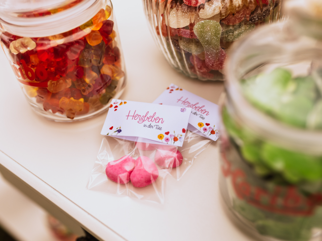 Candy-Bar Hochzeit Idee: Zwei kleine Klarsichtbeutel, mit jeweils zwei HARIBO Schaumzuckerherzen in Herzform. Auf einem Etikett steht „Herzbeben in der Tüte”. Im Hintergrund sind Glasbehälter mit HARIBO Süßigkeiten zu sehen.