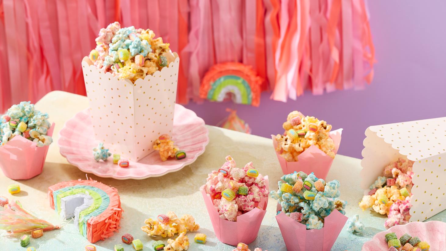 Einhorn Party Essen: Buntes Einhorn-Popcorn in Rosa, Blau und Gelb, serviert in rosa Tulpen-Förmchen und einer weiß-goldenen Popcorn-Box auf muschelförmigen Tellern. Das Popcorn ist getoppt mit HARIBO Pico-Balla, Pico-Balla Sauer, Rainbow, Pixel, Balla-Balla und Chamallows Minis, die auch verstreut auf dem Tisch liegen. Im Hintergrund eine Regenbogen-Piñata, rosa Fransen-Girlande und eine lila Wand – ein kreativer Snack für den Kindergeburtstag.