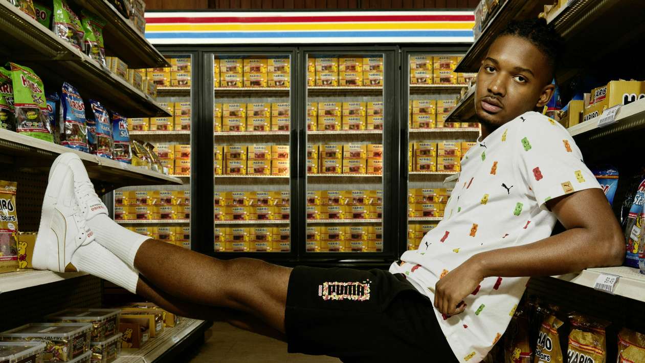Model sitzt gekleidet in T-Shirt, Shorts und Sneaker der PUMA x HARIBO Kollektion zwischen zwei Süßwarenregalen im Supermarkt