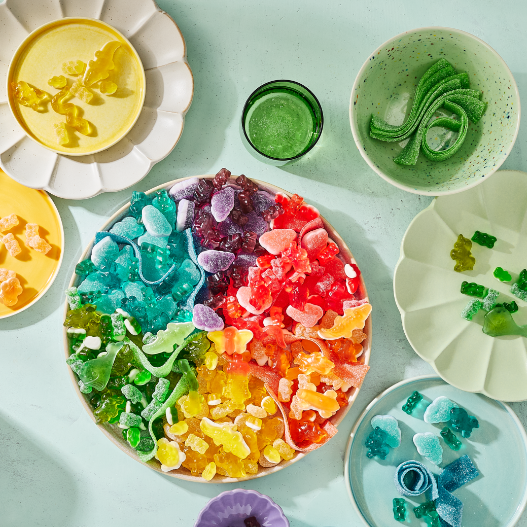 Pastel tabletop with a rainbow candy board piled with color‑sorted gummies—bears, hearts and ribbons—a playful unicorn snack station and easy favors for a unicorn birthday party.