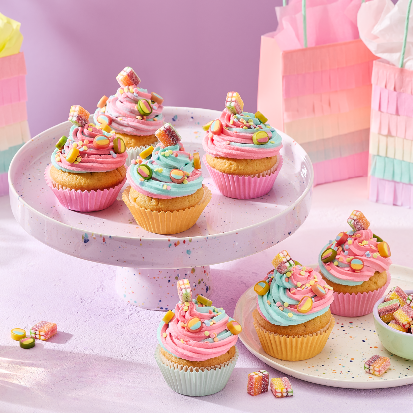 Cupcakes mit rosa und türkisem Frosting in pastellfarbenen Förmchen, dekoriert mit HARIBO Fruchtgummis, angerichtet auf einer gesprenkelten Etagere und einem passenden Teller. Daneben kleine Schälchen mit bunten Süßigkeiten, Gläser mit Getränken und regenbogenfarbene Geschenktüten im Fransen-Look vor einem lila Hintergrund.