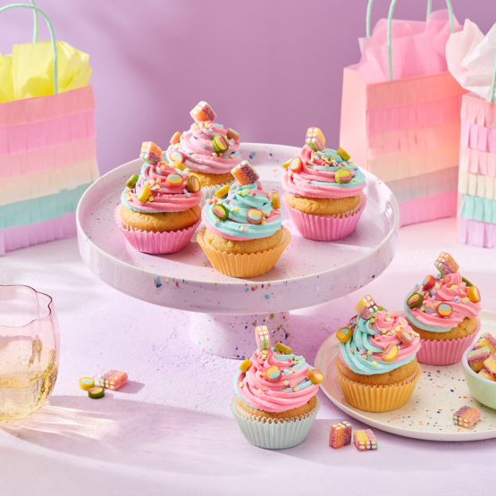 Cupcakes mit rosa und türkisem Frosting in pastellfarbenen Förmchen, dekoriert mit HARIBO Fruchtgummis, angerichtet auf einer gesprenkelten Etagere und einem passenden Teller. Daneben kleine Schälchen mit bunten Süßigkeiten, Gläser mit Getränken und regenbogenfarbene Geschenktüten im Fransen-Look vor einem lila Hintergrund.