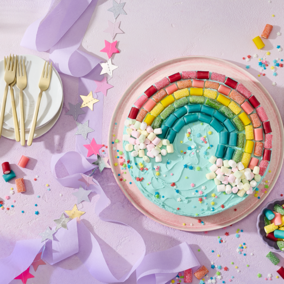 Ein runder Kuchen mit türkisem Frosting, dekoriert mit einem Regenbogen aus HARIBO Pico-Balla, Pico-Balla Sauer, Rainbow und Balla-Balla sowie bunten Pixel-Streuseln auf einem rosa Teller. Daneben Schälchen mit weiteren Fruchtgummis, goldene Gabeln auf weißen Tellern, gestreifte Servietten, eine Sternen-Girlande und lila Satinband auf einem hellflieder Hintergrund – ein farbenfroher Regenbogenkuchen für die Einhorn Party.