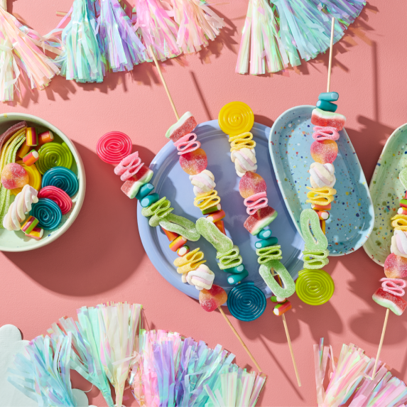 Einhorn Party Essen selber machen: Fünf farbenfrohe Regenbogen-Spieße bestückt mit HARIBO Balla-Balla, Bunte Schnecken, Wassermelonen, Pfirsiche, Apfelringe, Flower, Phantasia, Pasta Basta und Chamallows Twirlies auf pastellfarbenen Tellern. Daneben eine Schale mit weiteren Fruchtgummis. Dekoriert mit Quasten-Girlanden und Wolken-Servietten auf rosa Hintergrund.