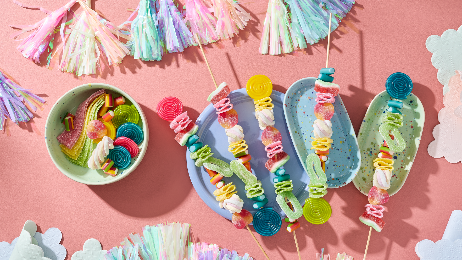Einhorn Party Essen selber machen: Fünf farbenfrohe Regenbogen-Spieße bestückt mit HARIBO Balla-Balla, Bunte Schnecken, Wassermelonen, Pfirsiche, Apfelringe, Flower, Phantasia, Pasta Basta und Chamallows Twirlies auf pastellfarbenen Tellern. Daneben eine Schale mit weiteren Fruchtgummis. Dekoriert mit Quasten-Girlanden und Wolken-Servietten auf rosa Hintergrund.