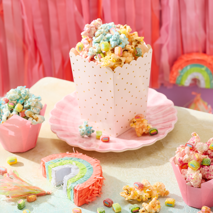Einhorn Party Essen: Buntes Einhorn-Popcorn in Rosa, Blau und Gelb, serviert in rosa Tulpen-Förmchen und einer weiß-goldenen Popcorn-Box auf muschelförmigen Tellern. Das Popcorn ist getoppt mit HARIBO Pico-Balla, Pico-Balla Sauer, Rainbow, Pixel, Balla-Balla und Chamallows Minis, die auch verstreut auf dem Tisch liegen. Im Hintergrund eine Regenbogen-Piñata, rosa Fransen-Girlande und eine lila Wand – ein kreativer Snack für den Kindergeburtstag.