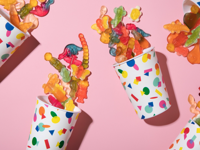 A variety of colorful candies and sweets displayed in glass jars and paper cones on a white table, including licorice wheels, gummy candies, and fruit-flavored treats. The text 'Snacks' is displayed in large yellow letters on a blue background, with the subtitle 'Sweet & homemade: Snacks for the birthday party.' This festive scene showcases delicious birthday party snacks and easy homemade snacks for birthday party setups — a sweet and essential part of how to plan a birthday party that guests of all ages will love.