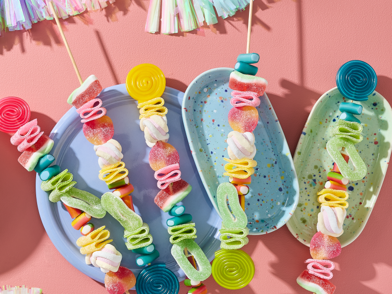 Einhorn Party Essen selber machen: Fünf farbenfrohe Regenbogen-Spieße bestückt mit HARIBO Balla-Balla, Bunte Schnecken, Wassermelonen, Pfirsiche, Apfelringe, Flower, Phantasia, Pasta Basta und Chamallows Twirlies auf pastellfarbenen Tellern. Daneben eine Schale mit weiteren Fruchtgummis. Dekoriert mit Quasten-Girlanden und Wolken-Servietten auf rosa Hintergrund.