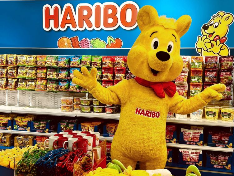 Der Goldbär steht vor einem Regal voller HARIBO-Produkte im HARIBO-Shop Wolfsburg.