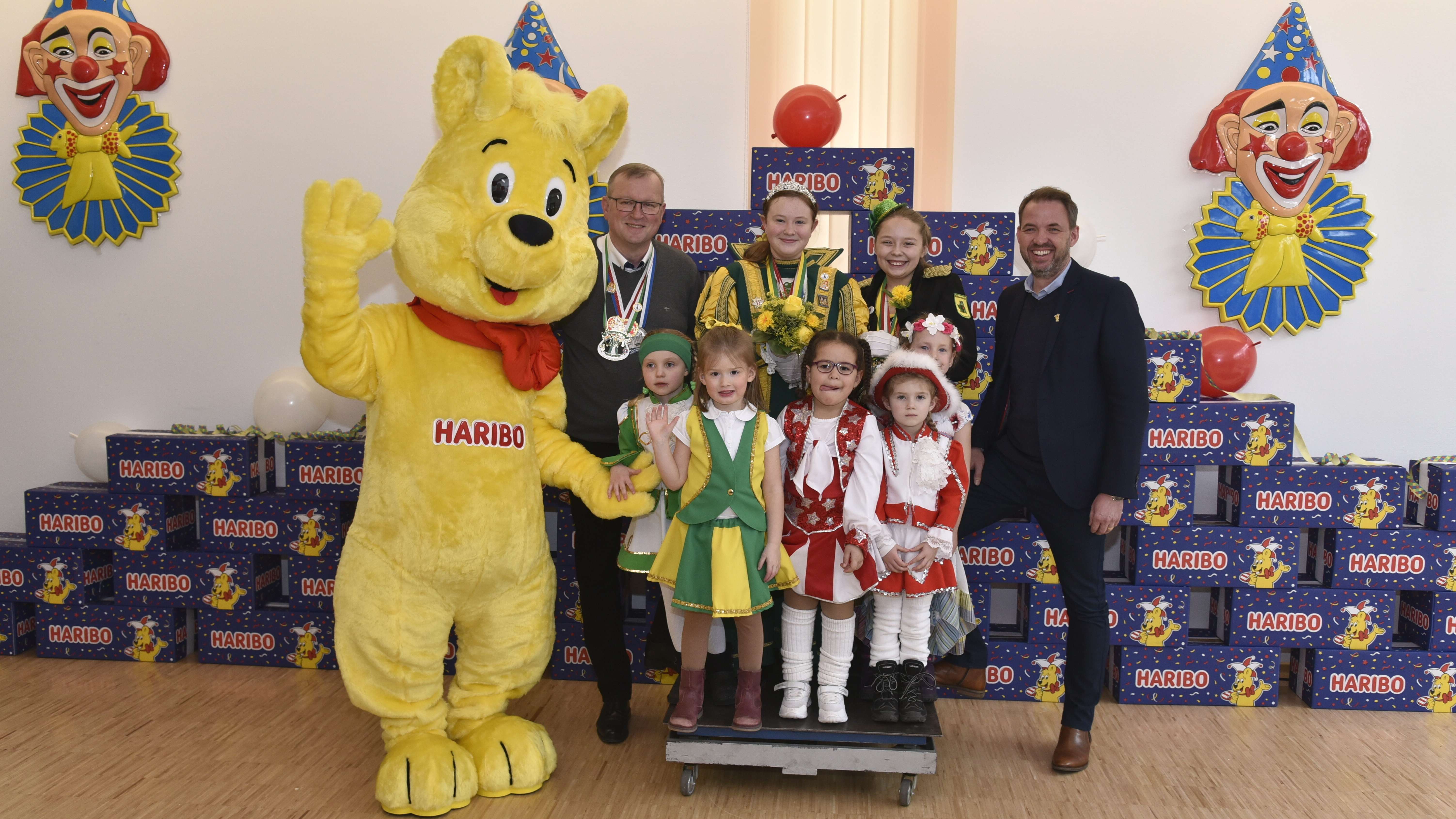 Winkender Goldbär mit Kindern in Karnevalskostümen auf einer Waage.
