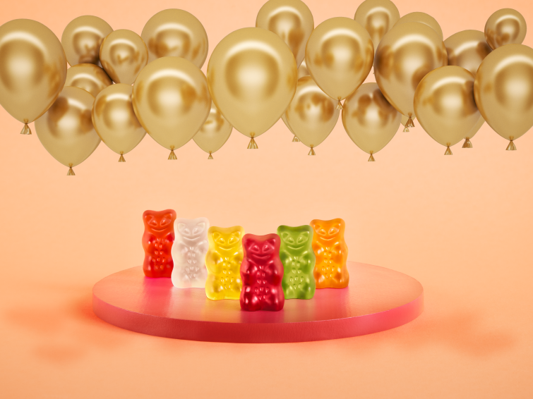 Sechs HARIBO Goldbären stehen auf einem Podest unter goldenen Luftballons