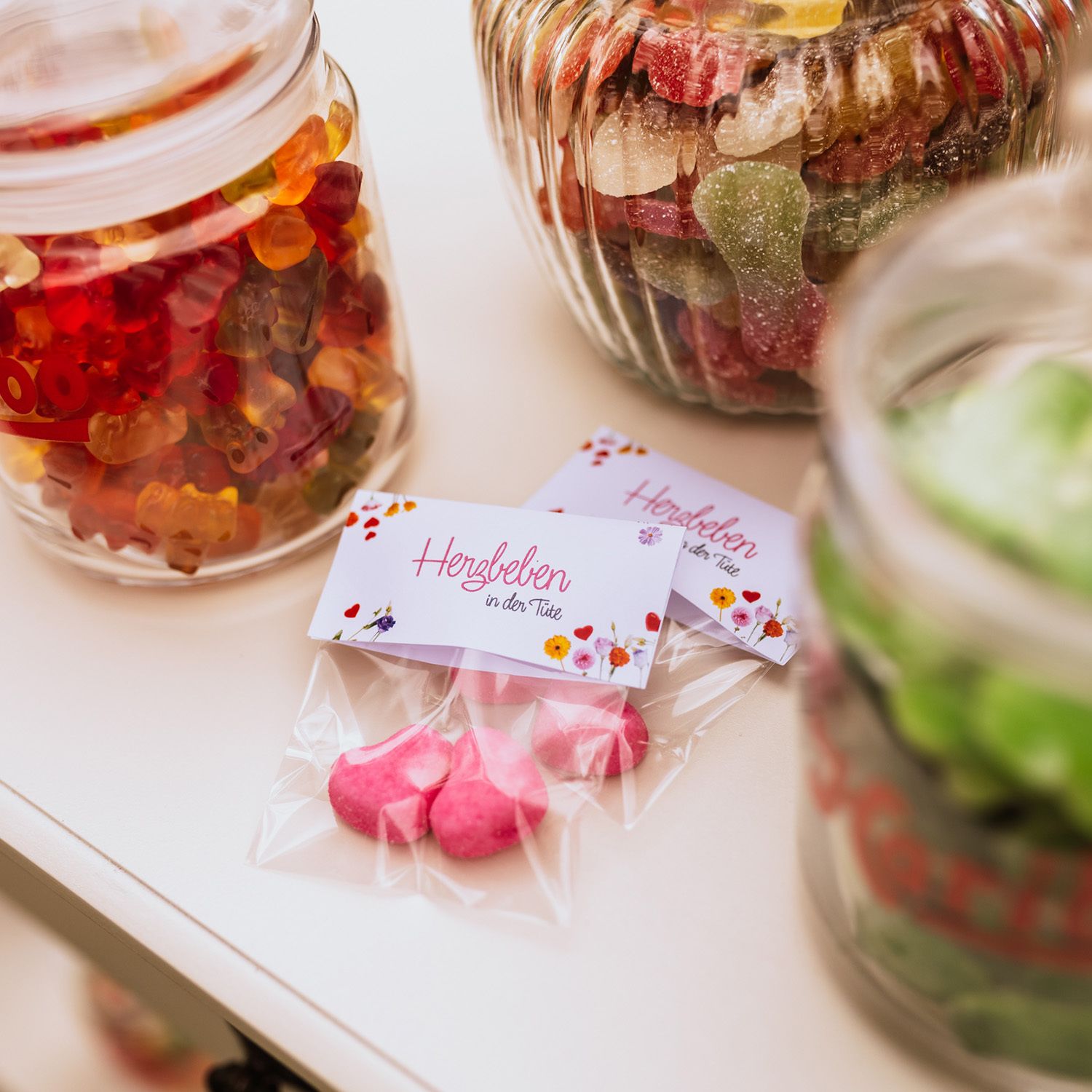 Candy-Bar Hochzeit Idee: Zwei kleine Klarsichtbeutel, mit jeweils zwei HARIBO Schaumzuckerherzen in Herzform. Auf einem Etikett steht „Herzbeben in der Tüte”. Im Hintergrund sind Glasbehälter mit HARIBO Süßigkeiten zu sehen.