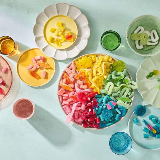 Eine großer Einhorn-Snackteller mit HARIBO Fruchtgummis, farblich nach Regenbogen sortiert von Rosa über Orange, Gelb und Grün bis Blau. Umgeben von kleineren Tellern und Schälchen in passenden Farben, auf denen die Süßigkeiten nach Farben getrennt angerichtet sind – darunter Bunte Schnecken, Balla-Balla und Pico-Balla. Farblich abgestimmte Gläser mit Getränken in Rosa, Bernstein, Grün und Blau ergänzen das Arrangement auf einem hellen Minz-Hintergrund – eine farbenfrohe Snack-Idee für die Geburtstagsparty.