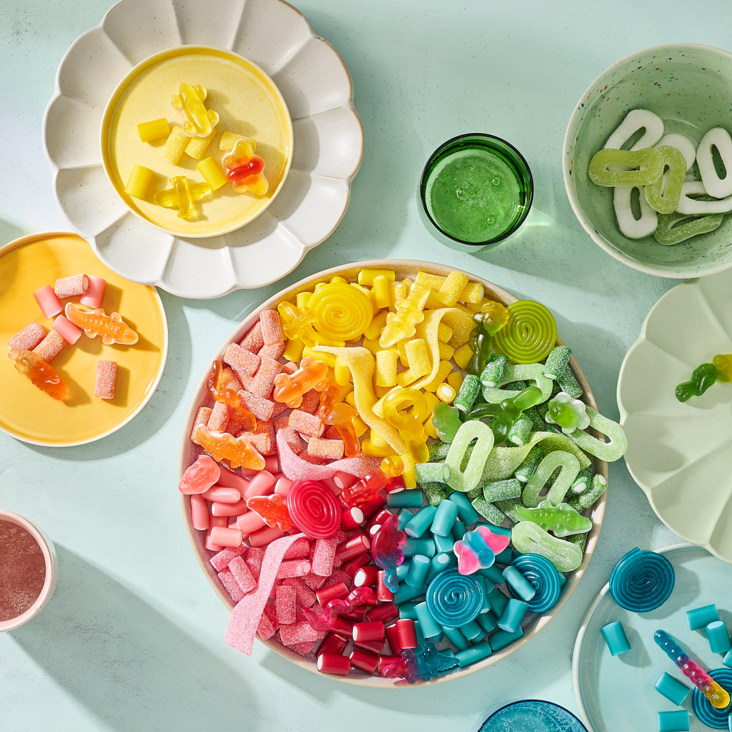 Eine großer Einhorn-Snackteller mit HARIBO Fruchtgummis, farblich nach Regenbogen sortiert von Rosa über Orange, Gelb und Grün bis Blau. Umgeben von kleineren Tellern und Schälchen in passenden Farben, auf denen die Süßigkeiten nach Farben getrennt angerichtet sind – darunter Bunte Schnecken, Balla-Balla und Pico-Balla. Farblich abgestimmte Gläser mit Getränken in Rosa, Bernstein, Grün und Blau ergänzen das Arrangement auf einem hellen Minz-Hintergrund – eine farbenfrohe Snack-Idee für die Geburtstagsparty.