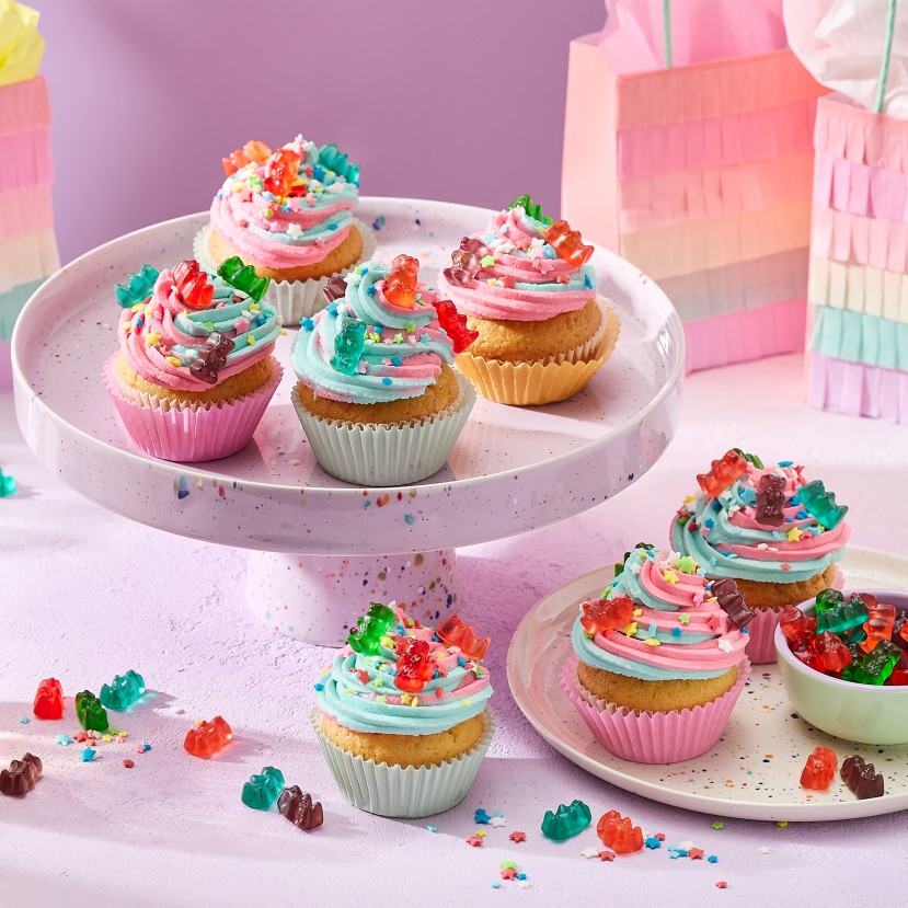 Cupcakes with pink and turquoise swirled frosting in pastel-colored cases, topped with HARIBO Goldbears and rainbow sprinkles, presented on a speckled cake stand and matching plate. Alongside are small bowls filled with more gummy bears, glasses with drinks and rainbow-colored fringed gift bags against a lilac background.
