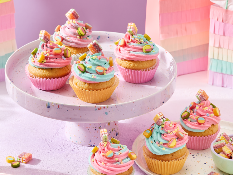 Cupcakes mit rosa und türkisem Frosting in pastellfarbenen Förmchen, dekoriert mit HARIBO Fruchtgummis, angerichtet auf einer gesprenkelten Etagere und einem passenden Teller. Daneben kleine Schälchen mit bunten Süßigkeiten, Gläser mit Getränken und regenbogenfarbene Geschenktüten im Fransen-Look vor einem lila Hintergrund.