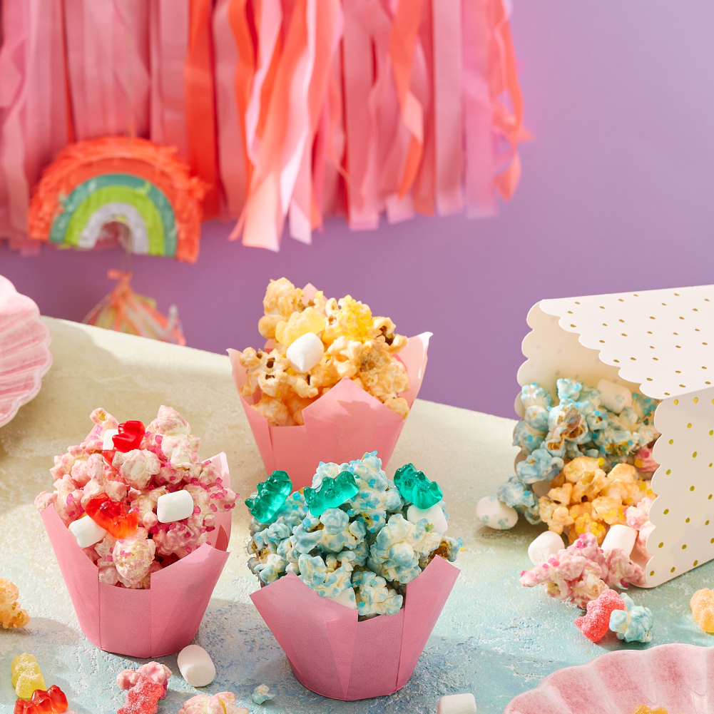 On a pastel party table, pink paper cups overflow with colorful unicorn popcorn in pastel colors, HARIBO Goldbears Wild Berry, and mini marshmallows—a sweet snack station and take‑home treat for a unicorn birthday party.
