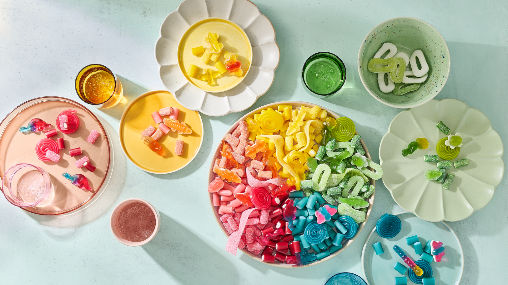 Eine großer Einhorn-Snackteller mit HARIBO Fruchtgummis, farblich nach Regenbogen sortiert von Rosa über Orange, Gelb und Grün bis Blau. Umgeben von kleineren Tellern und Schälchen in passenden Farben, auf denen die Süßigkeiten nach Farben getrennt angerichtet sind – darunter Bunte Schnecken, Balla-Balla und Pico-Balla. Farblich abgestimmte Gläser mit Getränken in Rosa, Bernstein, Grün und Blau ergänzen das Arrangement auf einem hellen Minz-Hintergrund – eine farbenfrohe Snack-Idee für die Geburtstagsparty.