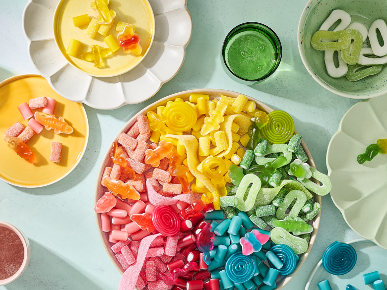 Eine großer Einhorn-Snackteller mit HARIBO Fruchtgummis, farblich nach Regenbogen sortiert von Rosa über Orange, Gelb und Grün bis Blau. Umgeben von kleineren Tellern und Schälchen in passenden Farben, auf denen die Süßigkeiten nach Farben getrennt angerichtet sind – darunter Bunte Schnecken, Balla-Balla und Pico-Balla. Farblich abgestimmte Gläser mit Getränken in Rosa, Bernstein, Grün und Blau ergänzen das Arrangement auf einem hellen Minz-Hintergrund – eine farbenfrohe Snack-Idee für die Geburtstagsparty.