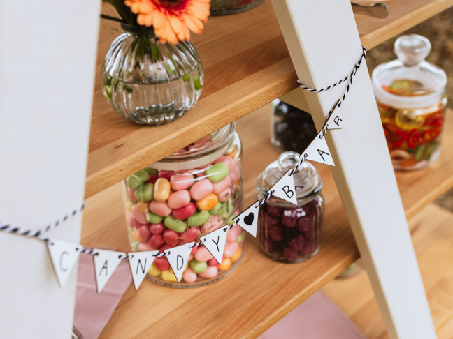 Candy-Bar zum Geburtstag oder Kindergeburtstag, gefüllt mit einer Vielzahl von HARIBO-Süßigkeiten in dekorativen Vorratsgläsern. Dazwischen: Blumen in Vasen. Im Vordergrund hängt eine kleine, dekorative Girlande mit den Worten „Candy Bar“.