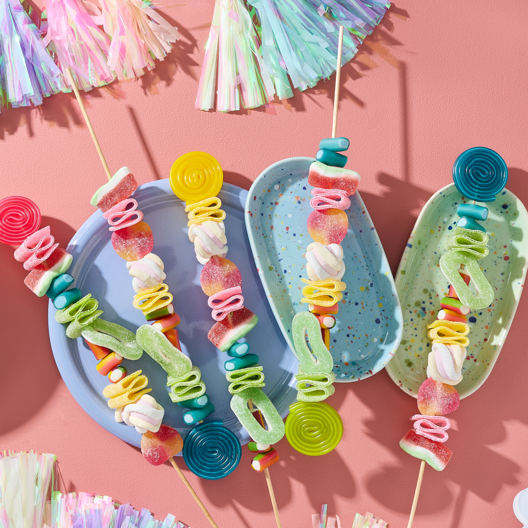 Einhorn Party Essen selber machen: Fünf farbenfrohe Regenbogen-Spieße bestückt mit HARIBO Balla-Balla, Bunte Schnecken, Wassermelonen, Pfirsiche, Apfelringe, Flower, Phantasia, Pasta Basta und Chamallows Twirlies auf pastellfarbenen Tellern. Daneben eine Schale mit weiteren Fruchtgummis. Dekoriert mit Quasten-Girlanden und Wolken-Servietten auf rosa Hintergrund.