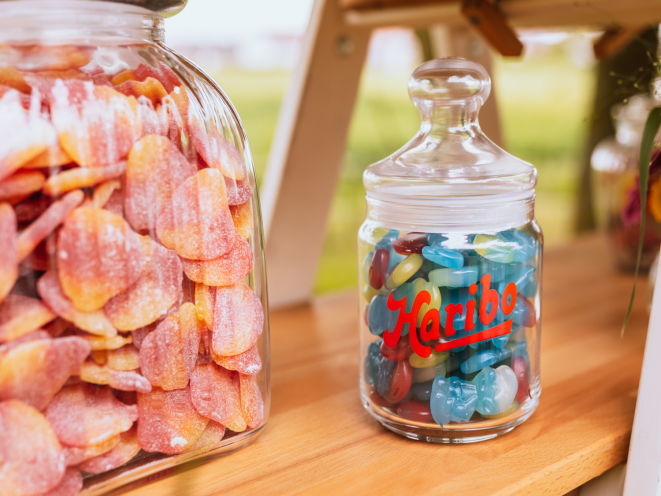 Two elegant glass jars displayed on a wooden table — one large jar filled with HARIBO Peaches and a smaller HARIBO-branded jar set against a bright outdoor backdrop. The text 'Bright Inspiration' is displayed in large yellow letters on a blue background, with the subtitle 'Candybar ideas: Bright inspiration for your party.' This vibrant setup showcases beautiful candy bar ideas with eye-catching candy for candy tables, perfect for creating a stunning pink candy buffet or mixing colors for a personalized candy table ideas display at any celebration.