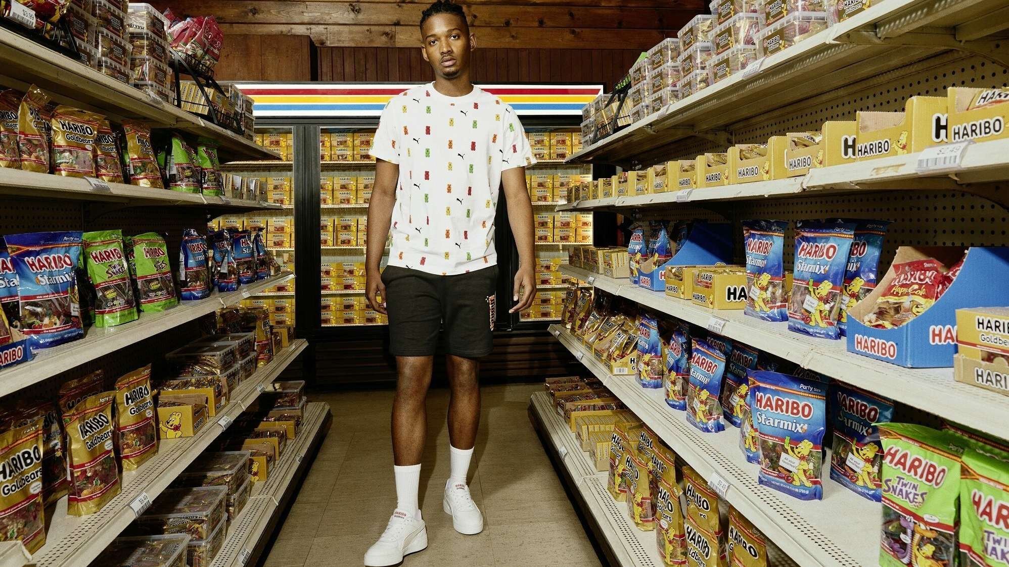 Model steht gekleidet in T-Shirt, Shorts und Schuhen der PUMA x HARIBO Kollektion zwischen zwei Süßwarenregalen im Supermarkt