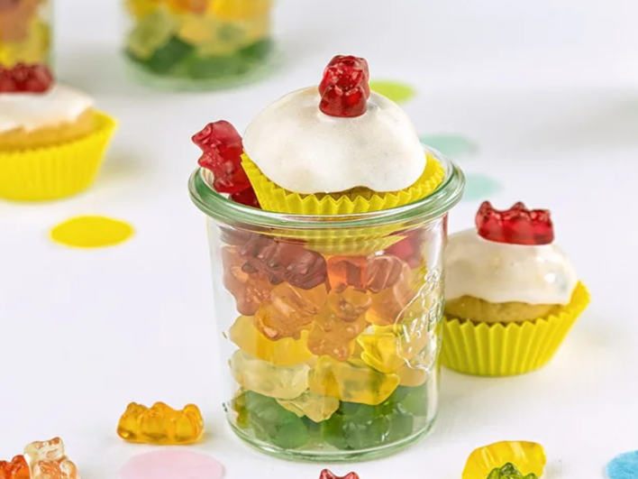 Creative cupcakes with white frosting topped with red HARIBO Goldbears, sitting on glass jars filled with colorful Goldbear candies — a delightful diy candy bar setup on a white table with confetti. The text 'Candybar for your birthday' is displayed in large yellow letters on a blue background, with the subtitle 'Candy bar for your birthday: Big fun for everyone.' This playful arrangement is the perfect inspiration for a candy bar for birthday party, showcasing fun and easy candy buffet ideas with colorful candy for candy tables that bring joy to any birthday party candy bar ideas display.