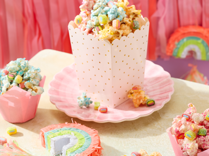 Einhorn Party Essen: Buntes Einhorn-Popcorn in Rosa, Blau und Gelb, serviert in rosa Tulpen-Förmchen und einer weiß-goldenen Popcorn-Box auf muschelförmigen Tellern. Das Popcorn ist getoppt mit HARIBO Pico-Balla, Pico-Balla Sauer, Rainbow, Pixel, Balla-Balla und Chamallows Minis, die auch verstreut auf dem Tisch liegen. Im Hintergrund eine Regenbogen-Piñata, rosa Fransen-Girlande und eine lila Wand – ein kreativer Snack für den Kindergeburtstag.